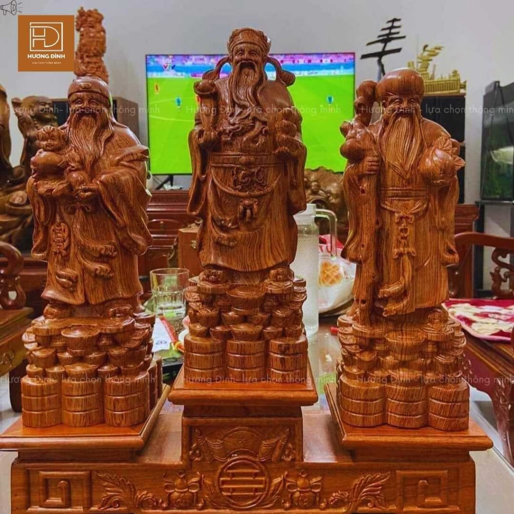tượng 3 ông Phúc Lộc Thọ được đặt ở trên mặt bàn. Đằng sau có một cái ti vi đang bật chương trình bóng đá