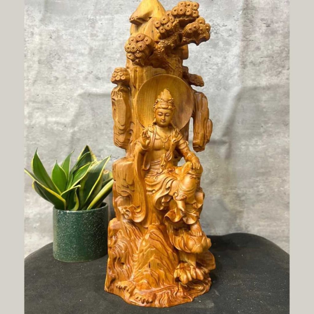 tượng Phật Quan âm tự tại làm từ gỗ. Ngài ngồi ở trên một gốc cây, trên đầu có một tán cây đang che nắng. Bên cạnh bức tượng có một cái cây kim tiền, chậu cây màu xanh, lá cây màu xanh lá cây và viền màu vàng.
